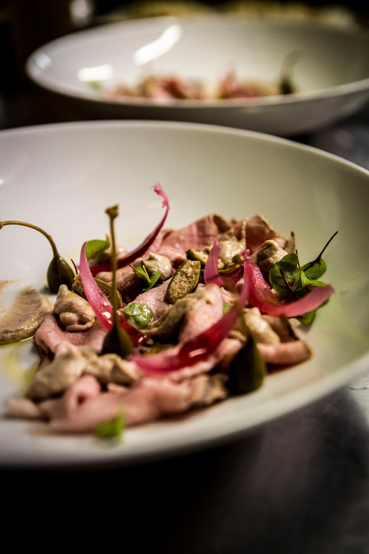 Assiette blanche de tranches de viande rosée avec câpres, oignons rouges marinés et feuilles vertes, sur une table sombre.