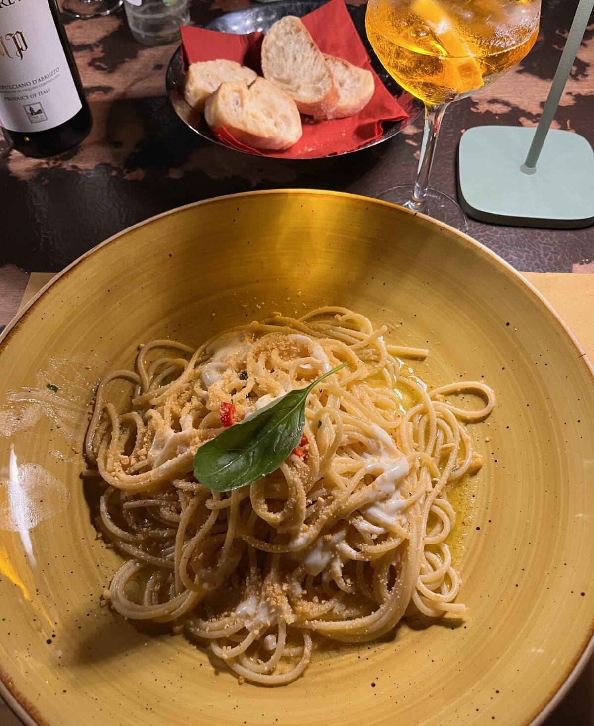 Teller mit Spaghetti in Käse-Pfeffer-Soße und Basilikumblatt, dazu Brot und ein Spritz-Glas auf einem Tisch