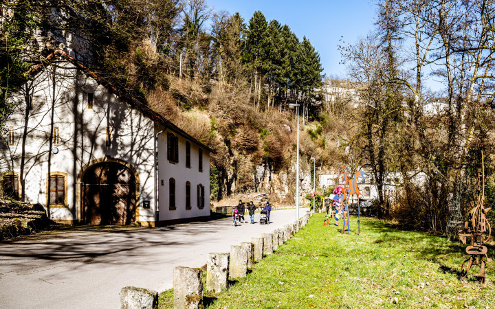 Walking path in the Hamm district, Luxembourg City, along the Alzette River and traditional houses, surrounded by greenery and cliffs.